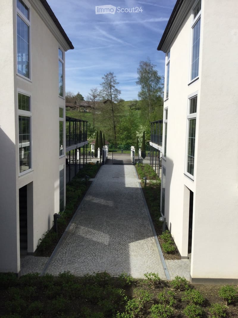 A paved pathway between two modern buildings, surrounded by greenery and small shrubs, leading to a gated entrance.