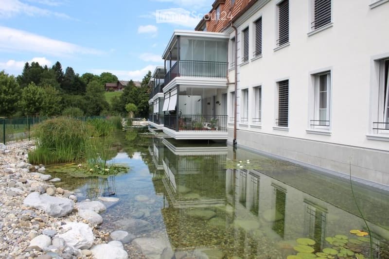 A modern building with a reflective glass balcony overlooking a serene water feature with lily pads and rocks, surrounded by greenery and trees.