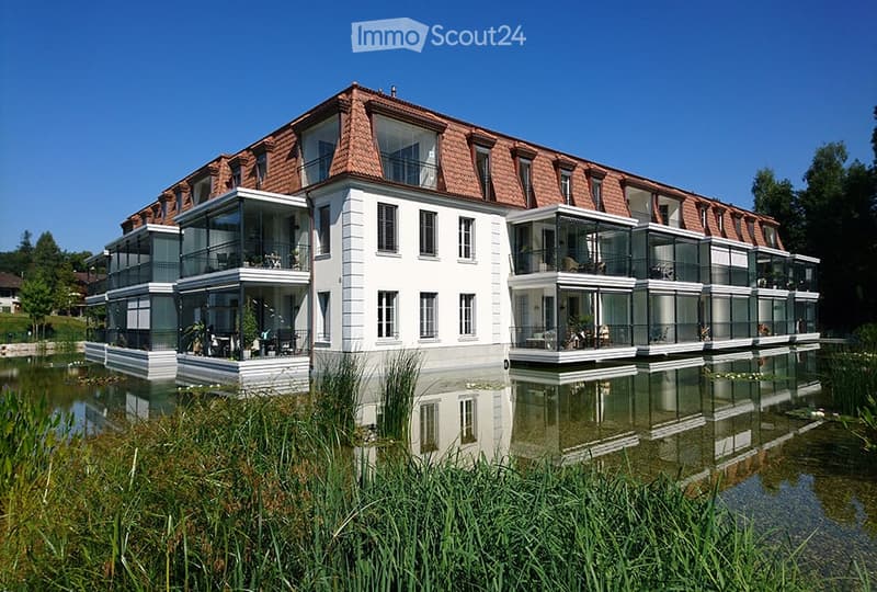 Modern two-story building with white walls, red-tiled roof, and large glass balconies overlooking a serene water body.