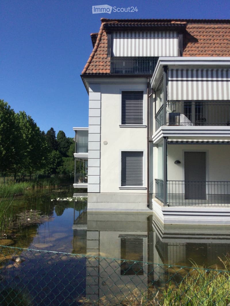 Modern two-story house with white walls, striped accents, and a red-tiled roof, situated on stilts above a small pond with a metal fence in the foreground.