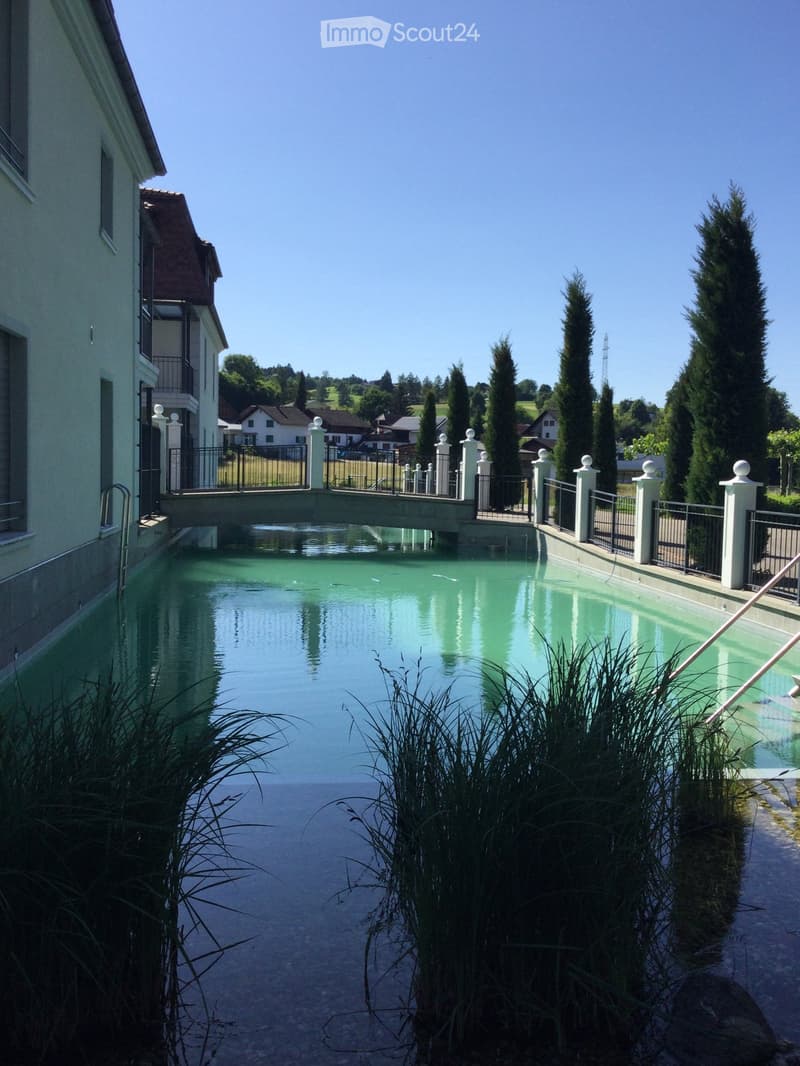 A serene swimming pool surrounded by a low fence, white balusters, and lush greenery, with a modern building in the background and a clear blue sky overhead.
