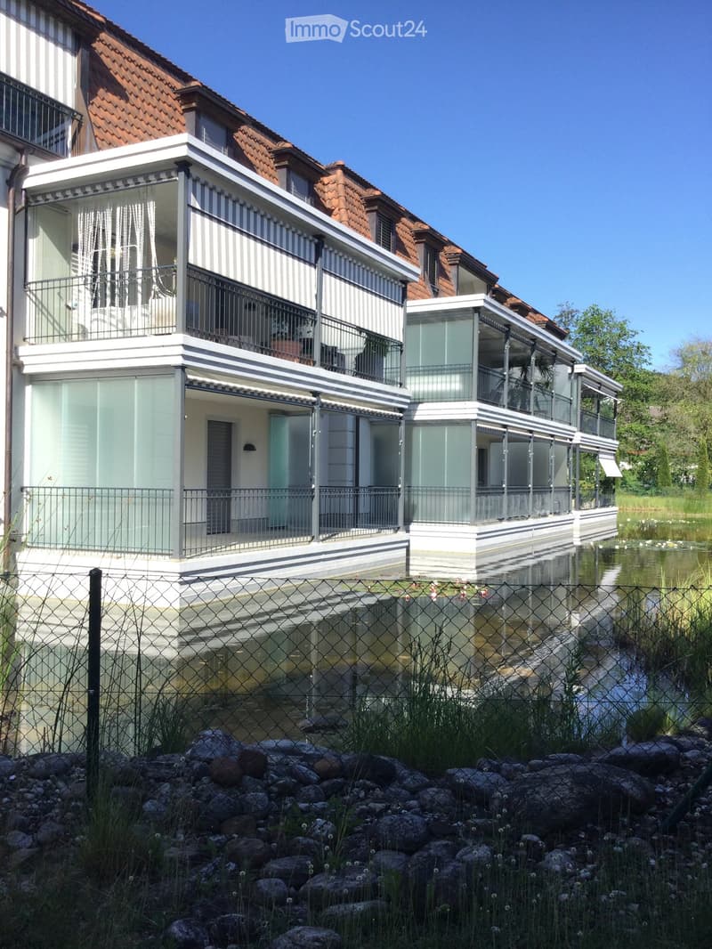 Modern two-story house with white and grey exterior, large glass balconies, and a red-tiled roof, situated near a water body with a fence and rocks in the foreground.