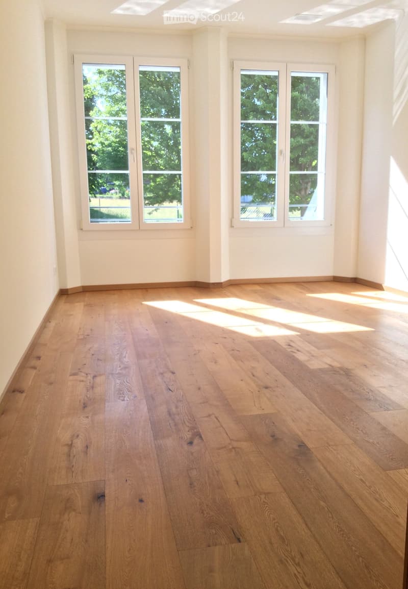 Empty room with light wooden flooring and large windows letting in natural light. The walls are painted a soft beige color, and the room has a clean, minimalist design with no furniture or decorations.