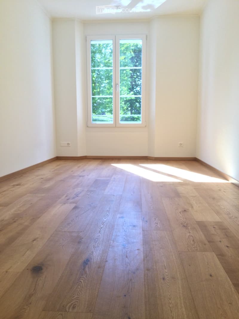 Empty room with light wooden flooring and white walls, featuring a large window with a view of greenery outside.