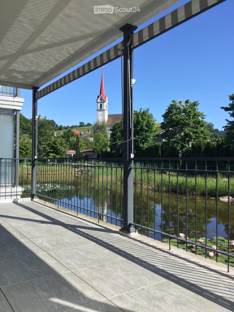 Balcony with a view of a lake, surrounded by a metal railing, featuring a covered awning and a serene landscape with trees and a church in the background.