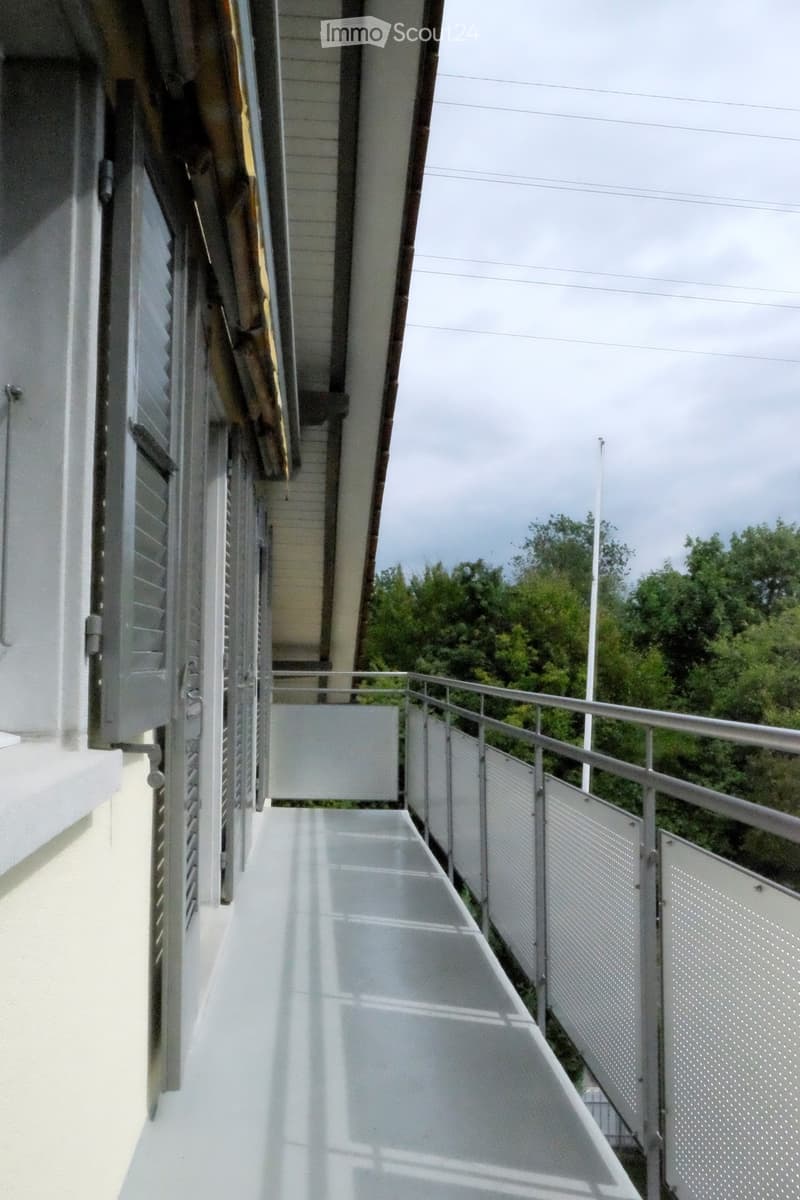 A modern balcony with white tiled flooring, metal railings, and a view of greenery in the background.