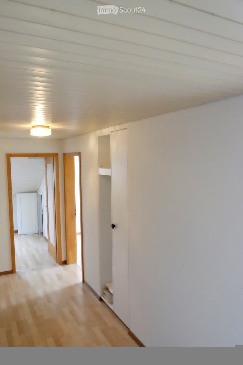 Empty hallway with light wooden flooring, white walls, and a built-in closet with a small shelf. The hallway has two open doors leading to other rooms.