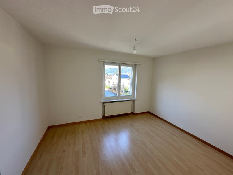 Empty room with wooden flooring, white walls, and a window offering a view of the outside. There is a radiator below the window and a light fixture on the ceiling.