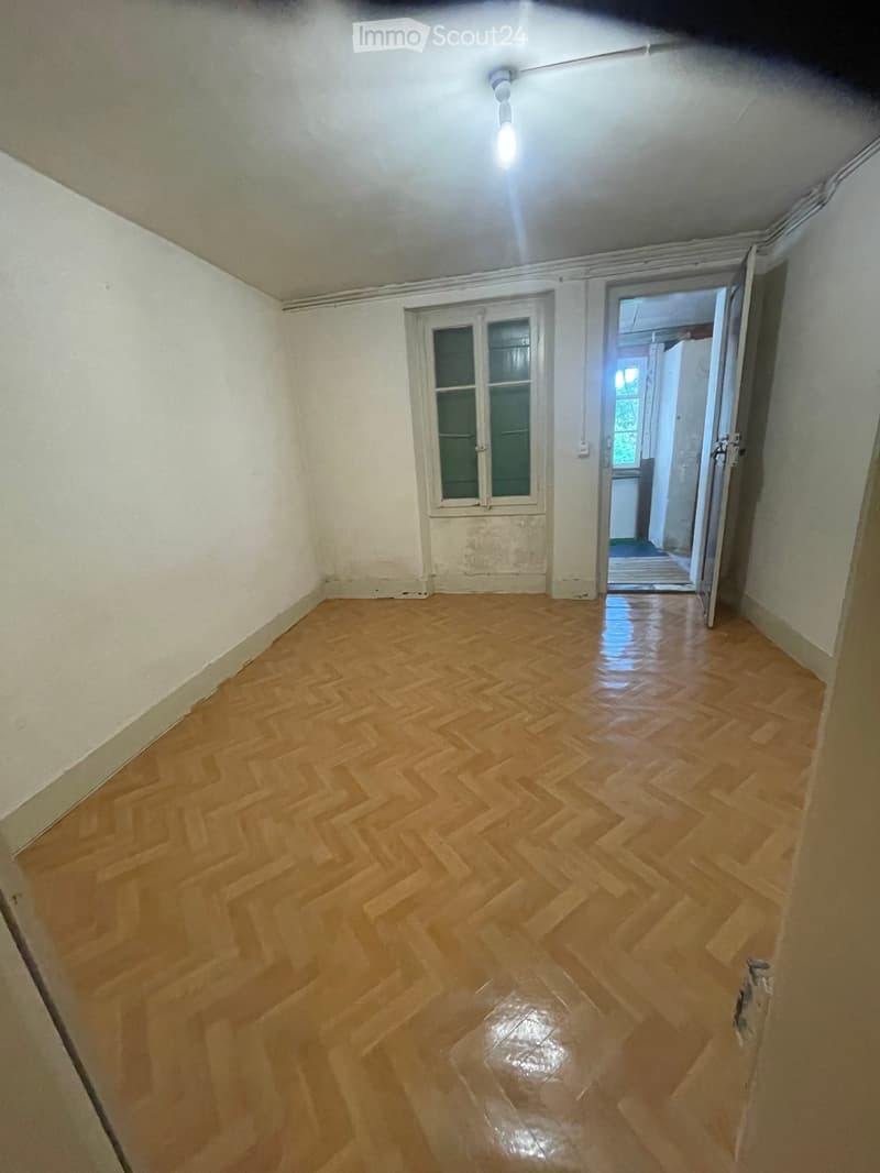 Empty room with wooden herringbone flooring, white walls, and a single window with frosted glass. An open doorway leads to another room.