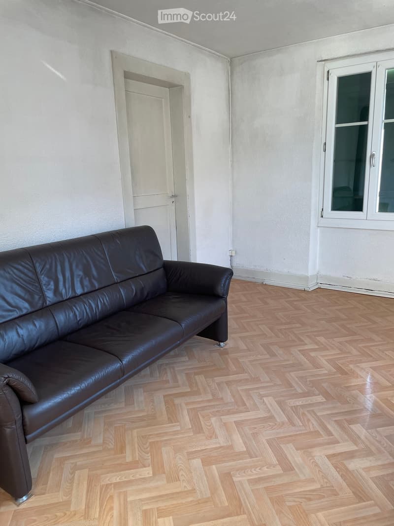 A furnished room with a dark brown leather sofa, wooden herringbone flooring, and a white door leading to another room.