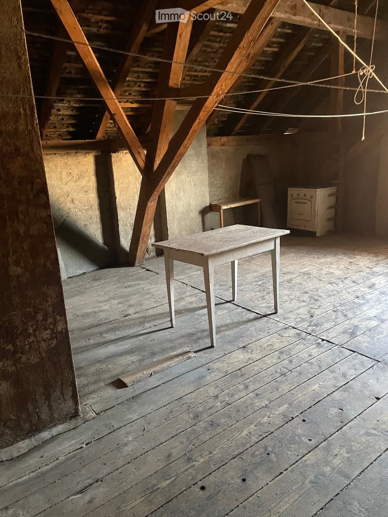 Empty attic space with wooden beams, exposed wooden floor, and minimal furnishings including a small table and a bench.
