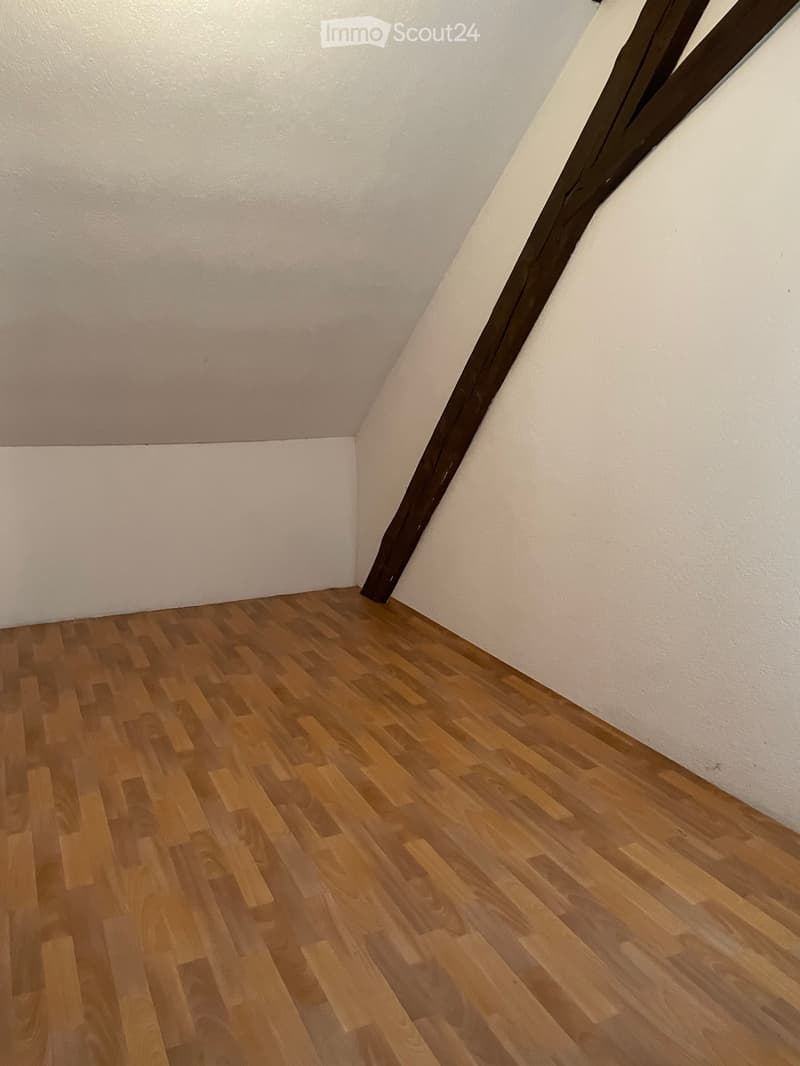 Empty attic room with wooden flooring and white walls, featuring exposed wooden beams and a slanted ceiling.