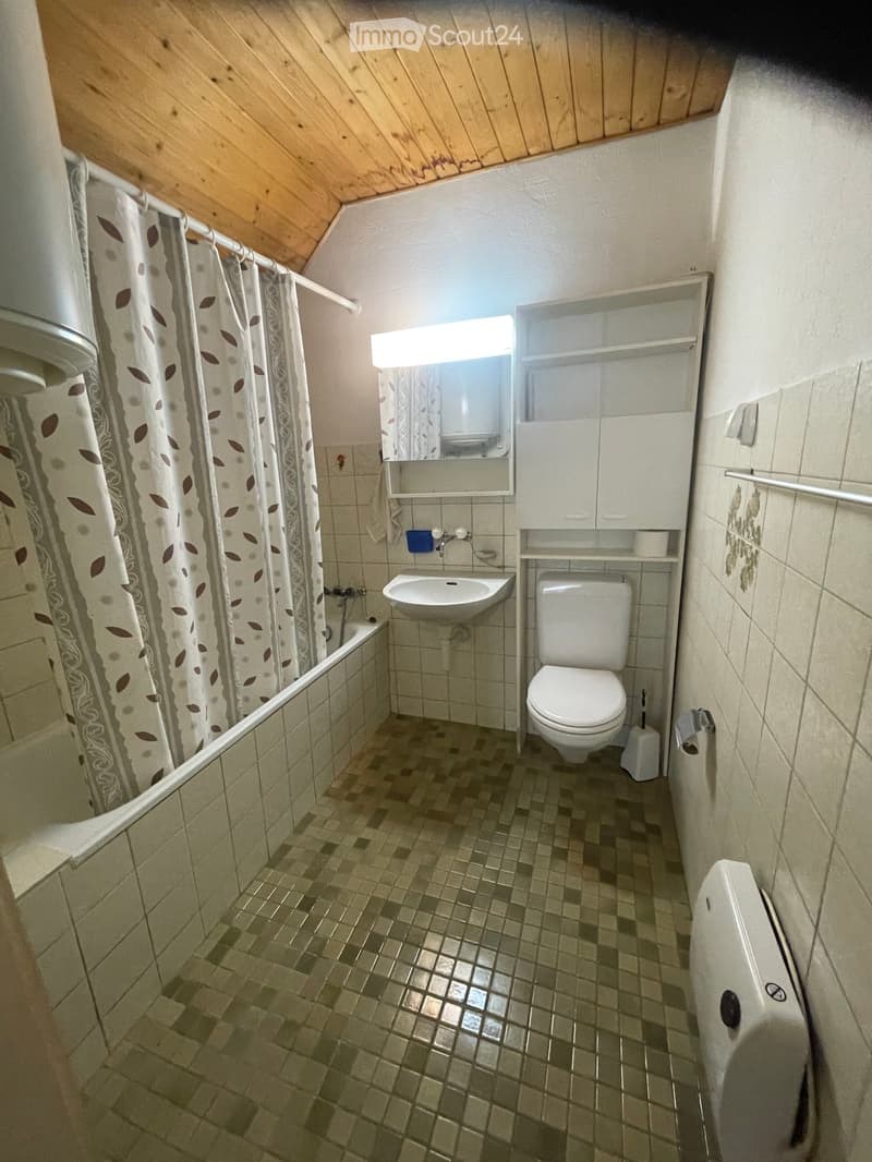 Small bathroom with a toilet, sink, bathtub, and mirror. The room features tiled walls and a wooden slanted ceiling.