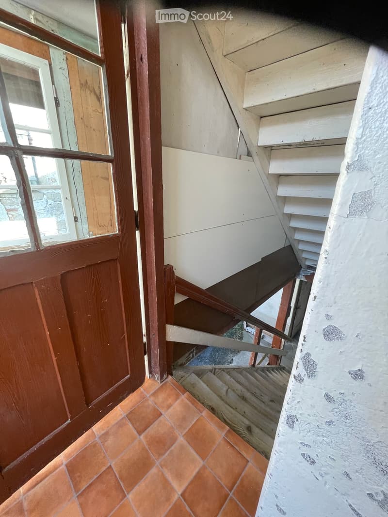 A narrow hallway with orange tiled floor, wooden door, and a staircase leading upwards.