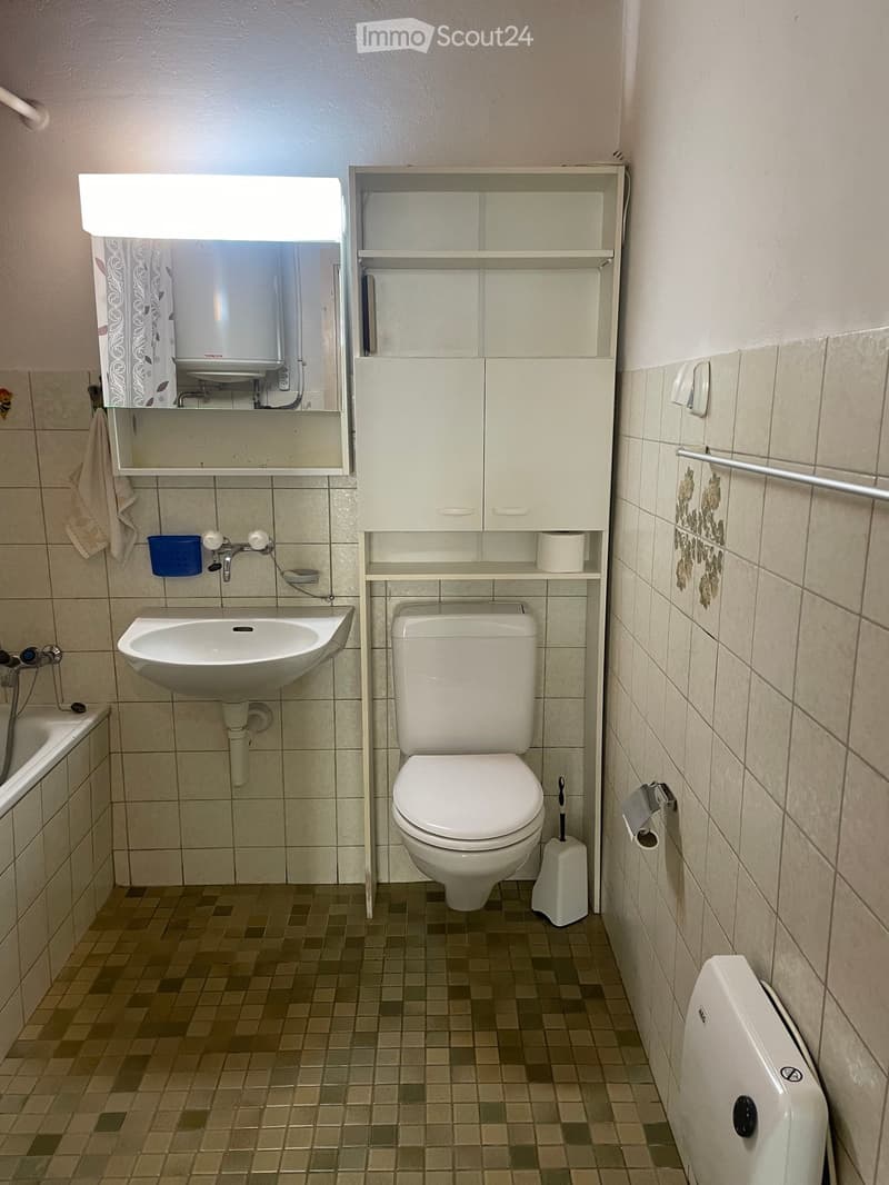 A small bathroom with a toilet, sink, mirror, and built-in cabinet. The walls are tiled, and the floor has small square tiles.