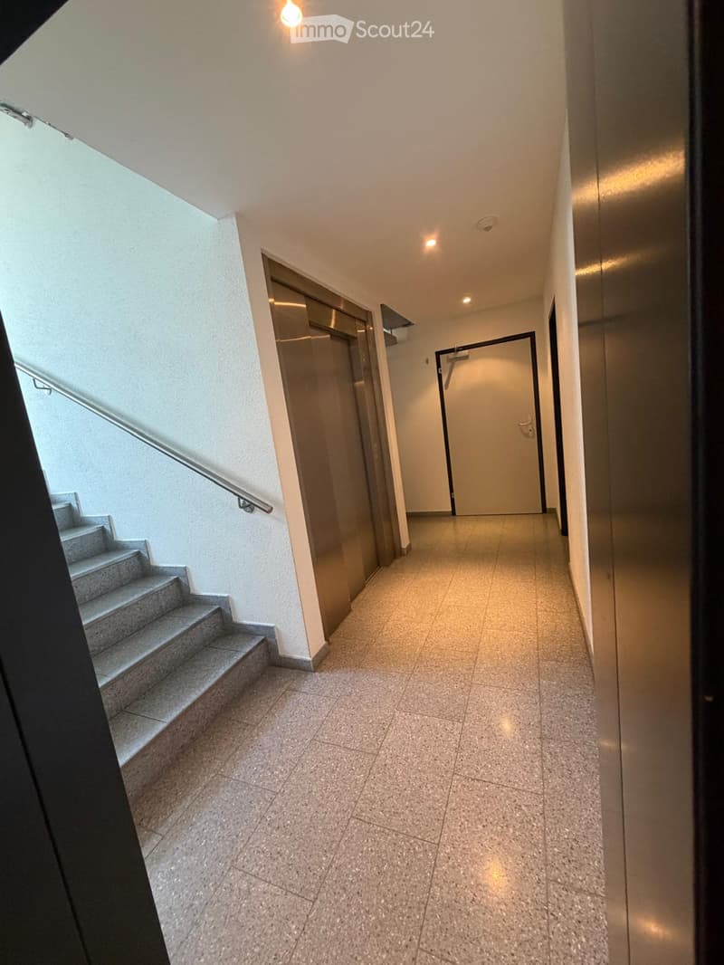 Clean hallway with tiled floor, staircase on the left, elevator on the right, and a closed door at the end.