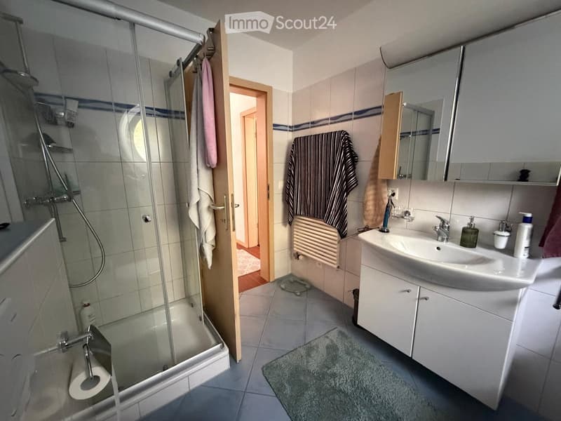 Modern bathroom with a glass-enclosed shower, a white sink with a mirror cabinet, and a toilet. Towels are hanging on the wall, and a small rug is placed on the floor.