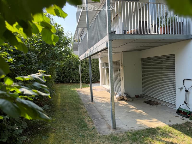 Modern outdoor area with a paved pathway, grassy lawn, and a covered balcony area with metal railings and potted plants.