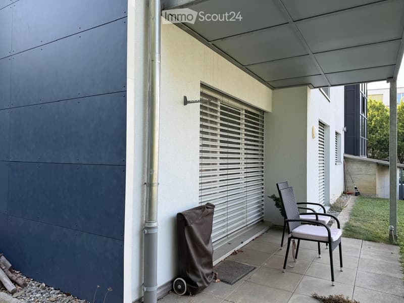 Covered outdoor area with tiled floor, grey wall, window with blinds, covered grill, and a single chair.