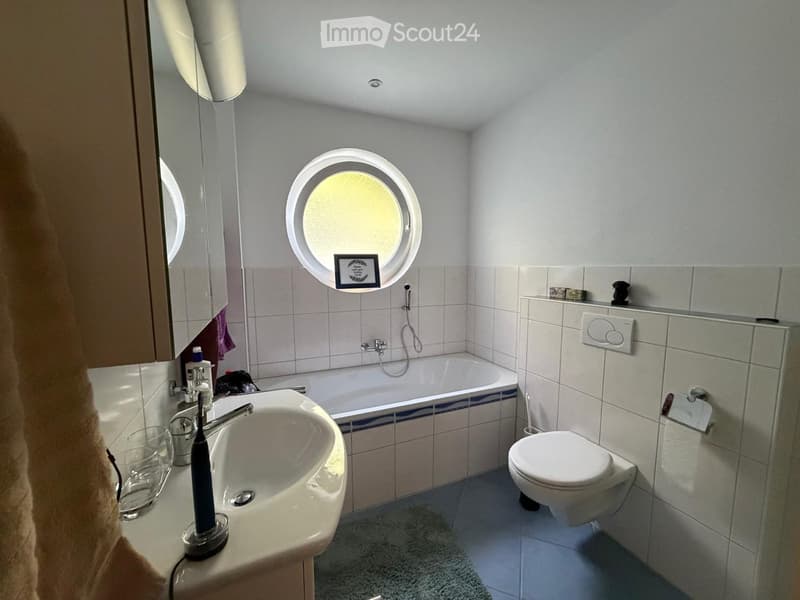 Modern bathroom with a sink, bathtub, toilet, and a large round window above the bathtub. Includes a mirror, toothbrush holder, and a towel on the side.