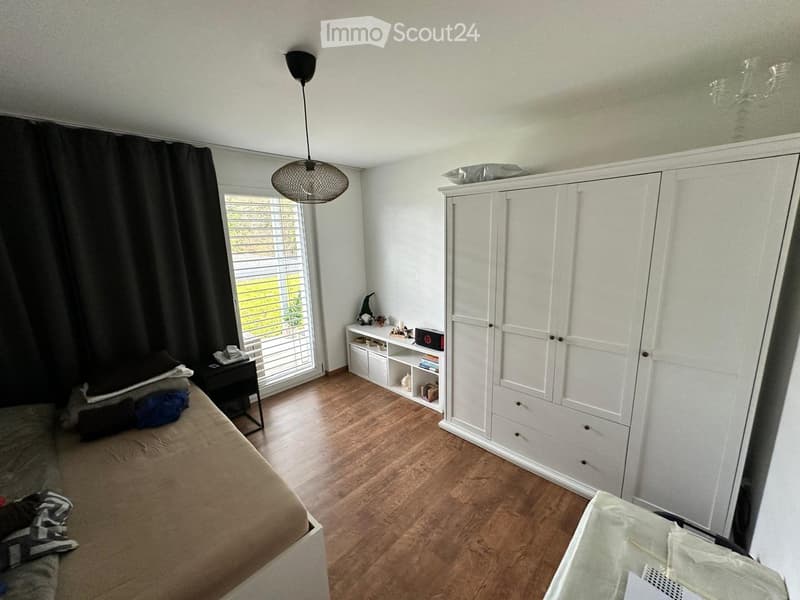 Well-lit bedroom with wooden flooring, a large bed, a white wardrobe, a nightstand, and a shelf with decorative items.