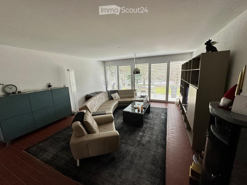 Modern living room with a curved sofa, coffee table, bookshelf, and fireplace. Large windows with blinds provide natural light.