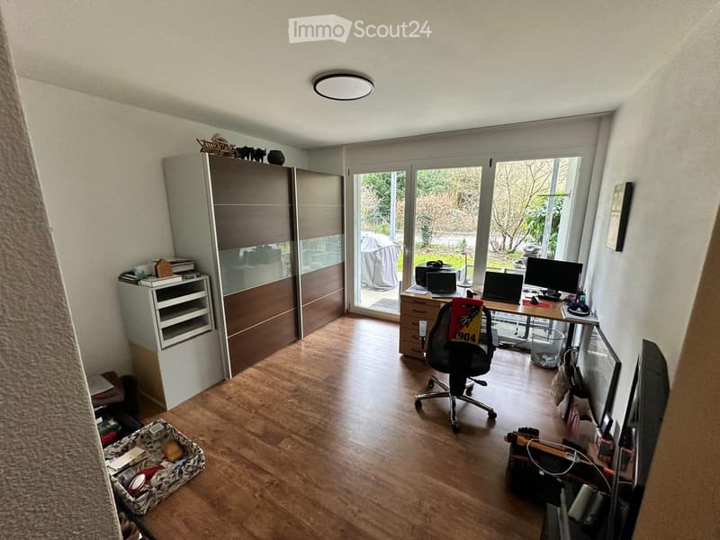 Well-lit room with wooden flooring, large sliding glass doors leading to a garden, a desk with multiple screens and laptops, a comfortable office chair, and a tall cabinet with drawers.