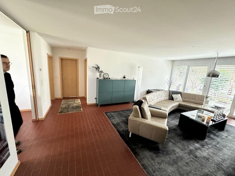 Modern living room with a beige sectional sofa, a black coffee table, a grey cabinet, and a clock on top. The room features large windows with blinds and a dark grey rug on the floor.