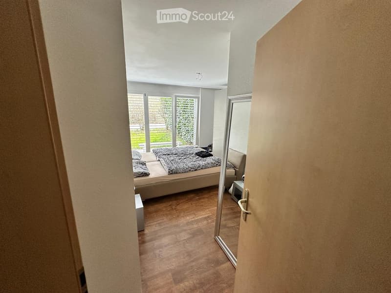 Modern bedroom with wooden flooring, large windows, a bed with patterned bedding, a nightstand, and a small sofa.