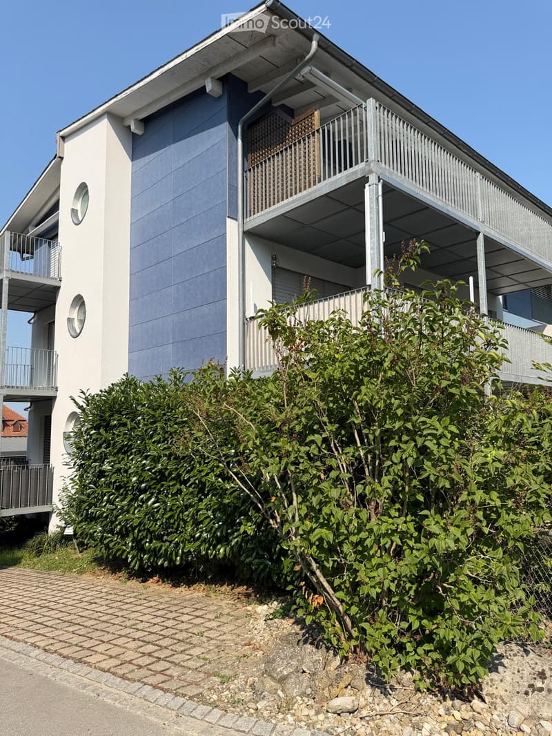 Modern two-story building with white and grey exterior, multiple balconies, and lush green bushes in front.