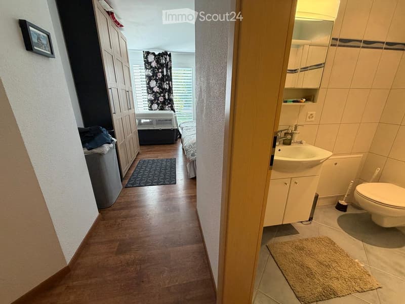 A narrow hallway with wooden flooring, leading to a bedroom and a bathroom. The hallway features a small rug, a pet carrier, and a partially visible bed with bedding.