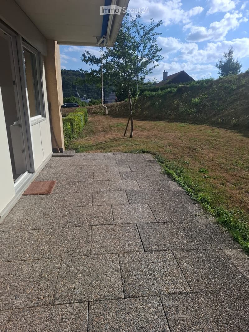 Outdoor patio area with stone tiles, small grassy patch, and a view of a hillside with trees and a house in the background.