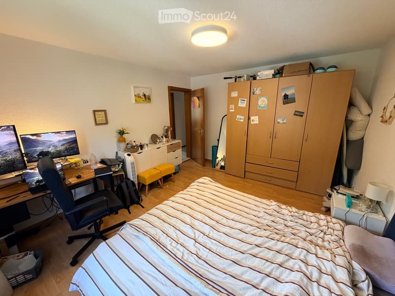 Well-lit bedroom with a bed, desk, chair, wardrobe, and decorative items. The room features wooden flooring and a modern aesthetic.