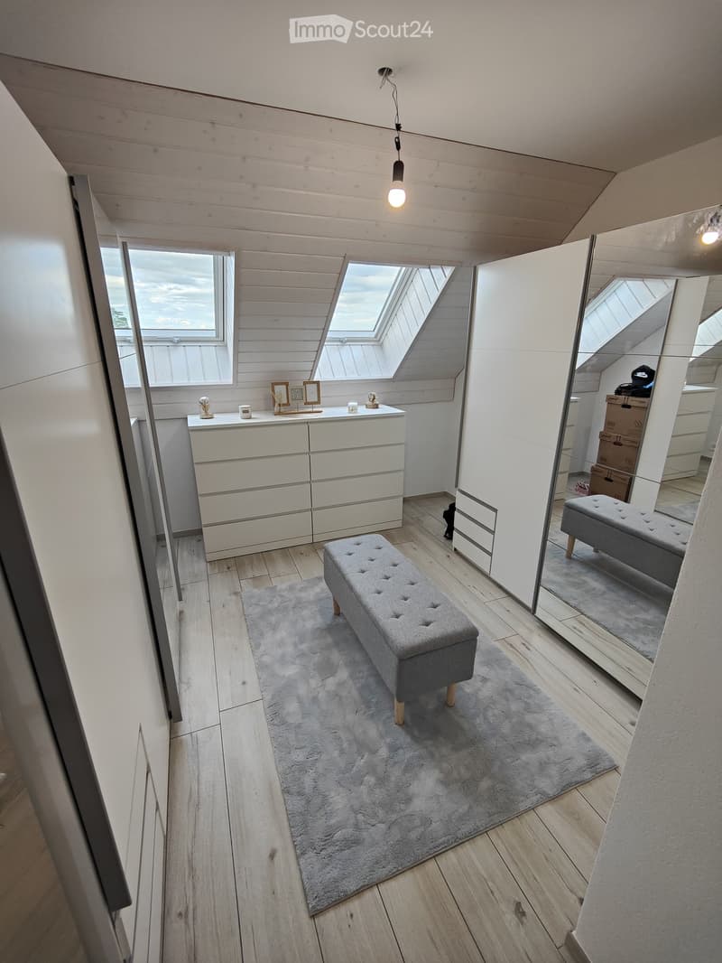 Well-lit room with wooden flooring, a grey bench on a rug, a white cabinet with drawers, a mirror, and decorative items on the cabinet.