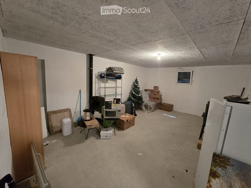 The room has a concrete ceiling and walls, with a table, chair, microwave, refrigerator, shelf, screen, stool, trash can, and a small Christmas tree.