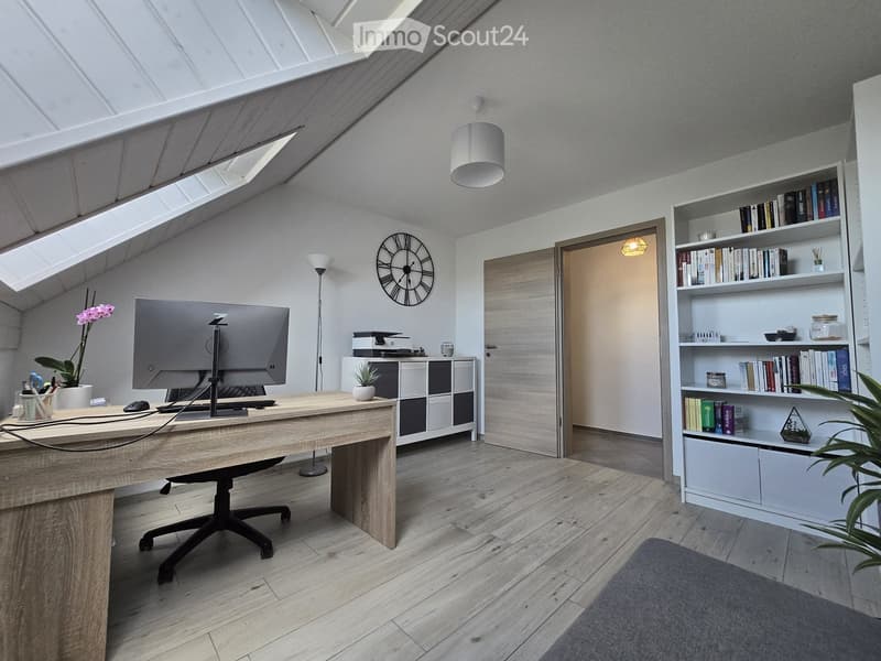 Modern, well-lit room with a wooden desk, ergonomic chair, large screen, bookshelf filled with books, decorative clock, potted plants, and a grey carpet.