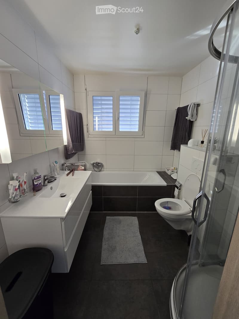 Modern bathroom with white tiles, a bathtub, a toilet, a sink with a mirror, and a glass-enclosed shower. The room features dark tiled flooring and a small gray rug in the center.