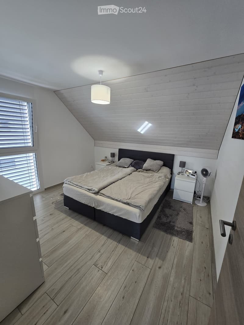 Modern bedroom with wooden flooring, slanted ceiling, large window with blinds, double bed with pillows and blankets, nightstand with lamp, fan, and picture frame.