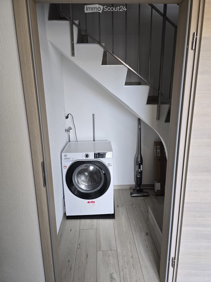 Modern bathroom with a white washing machine, a vacuum cleaner, and wooden flooring.