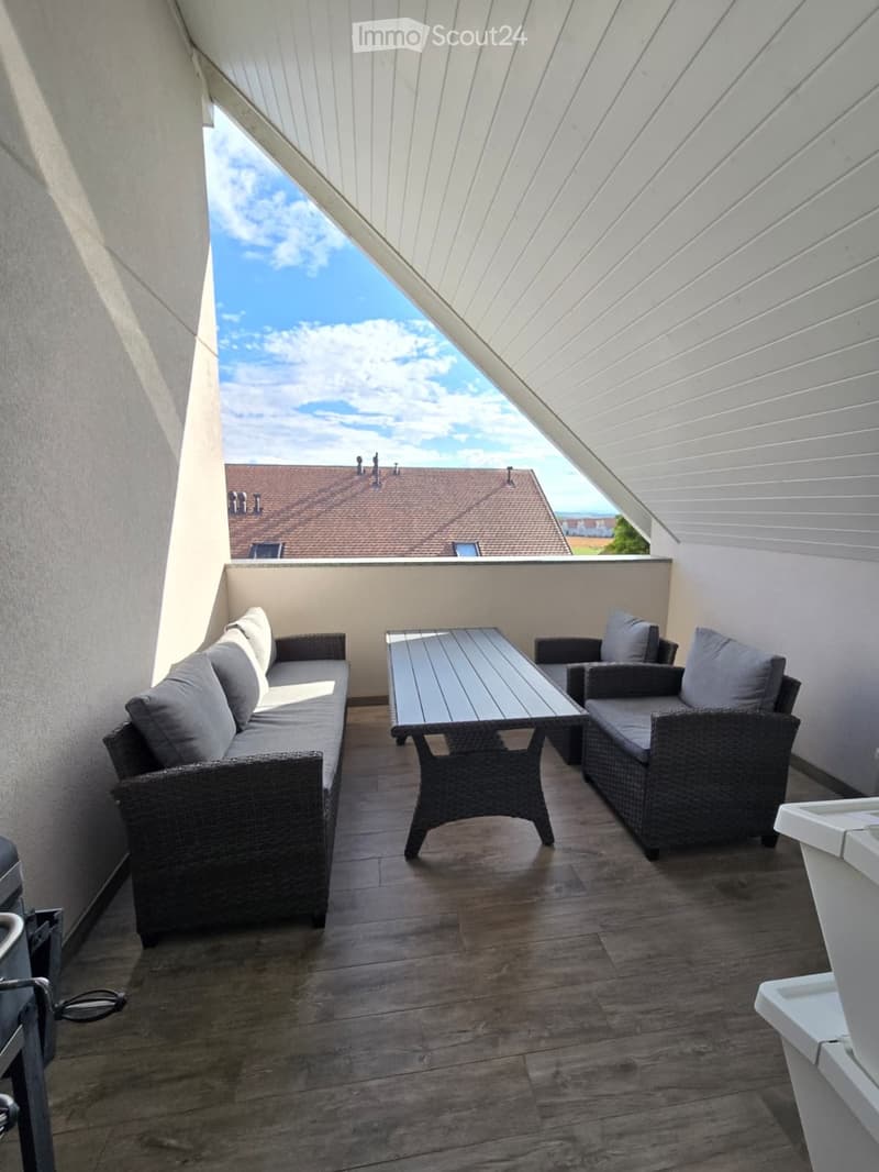 Balcony with wooden floor, white walls, grey wicker sofa, table, and chairs, overlooking a roofed building.