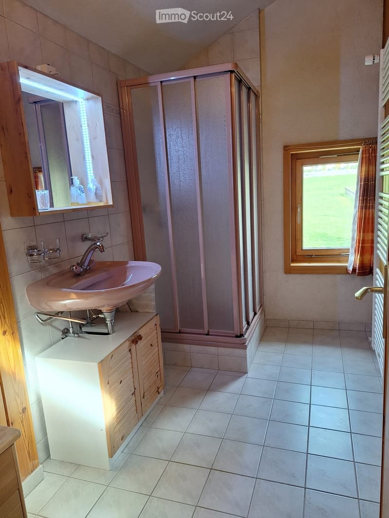 Bathroom with a pink sink, wooden cabinet, mirror with LED lighting, shower enclosure, tiled floor, and a window with a curtain.