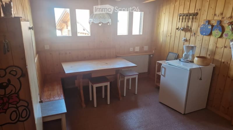 A cozy, wooden-paneled room with a dining table, two stools, a refrigerator, a laptop, a coffee maker, and a bowl on top. The room features a chandelier, a radiator, and a small cabinet with a decorative design on the door.