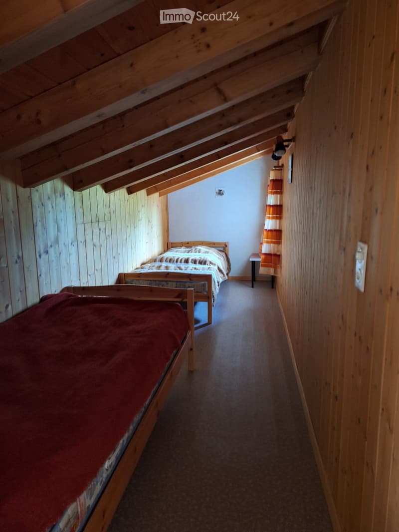Cozy attic bedroom with wooden walls and slanted ceilings, featuring two single beds, a small table, and a curtained window.