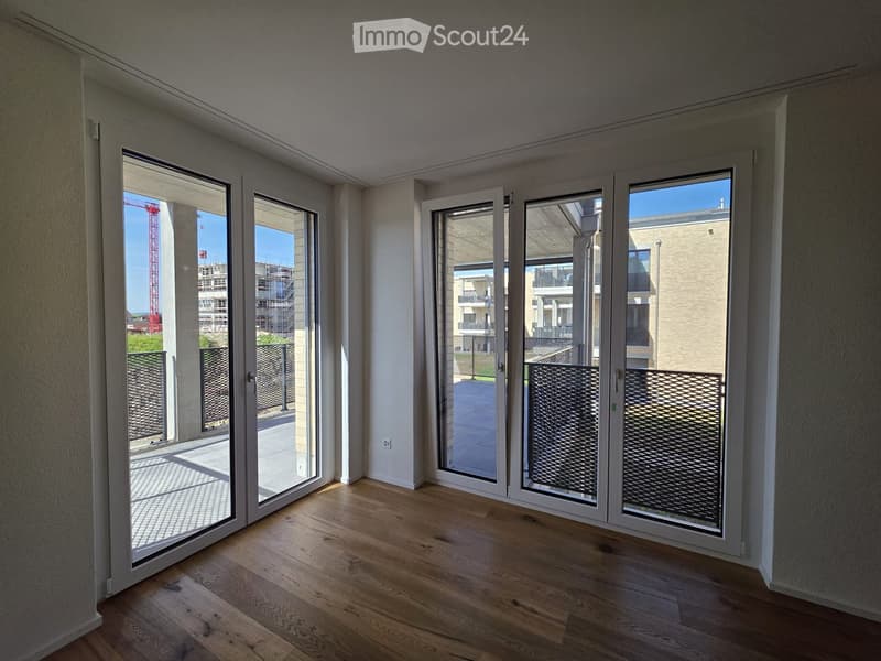 Empty room with wooden flooring and large sliding glass doors leading to a balcony area. The walls are painted white, and the space is well-lit with natural light.