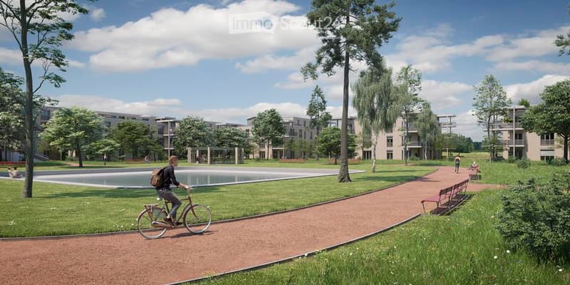 Garden / Outdoor Area. A park-like outdoor area with a paved pathway, grassy areas, trees, and modern apartment buildings in the background. A person is riding a bicycle along the pathway.
