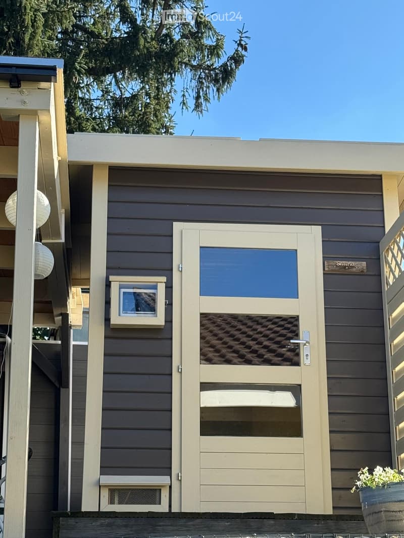 Exterior of small house, grey wooden cladding, white door, small box above door, window above door, plants in pots, hanging white lights