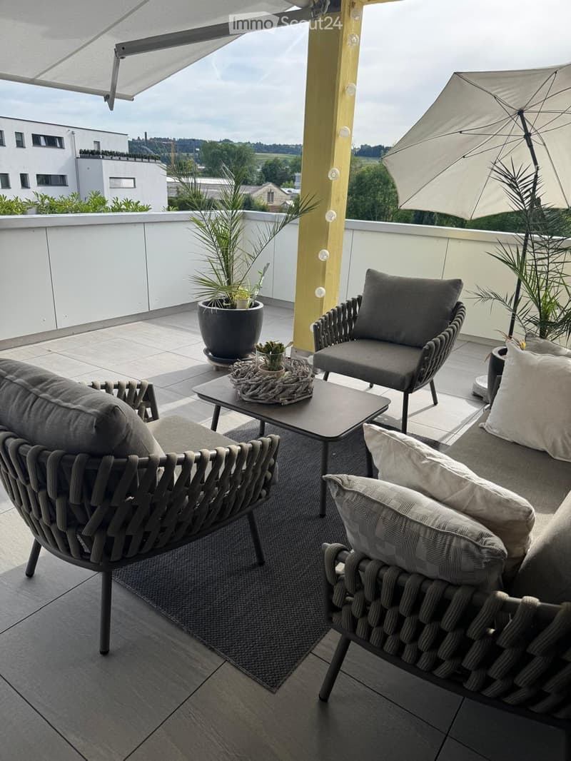 Modern balcony with grey wicker chairs, a coffee table, umbrella, and potted plants, overlooking a scenic view.