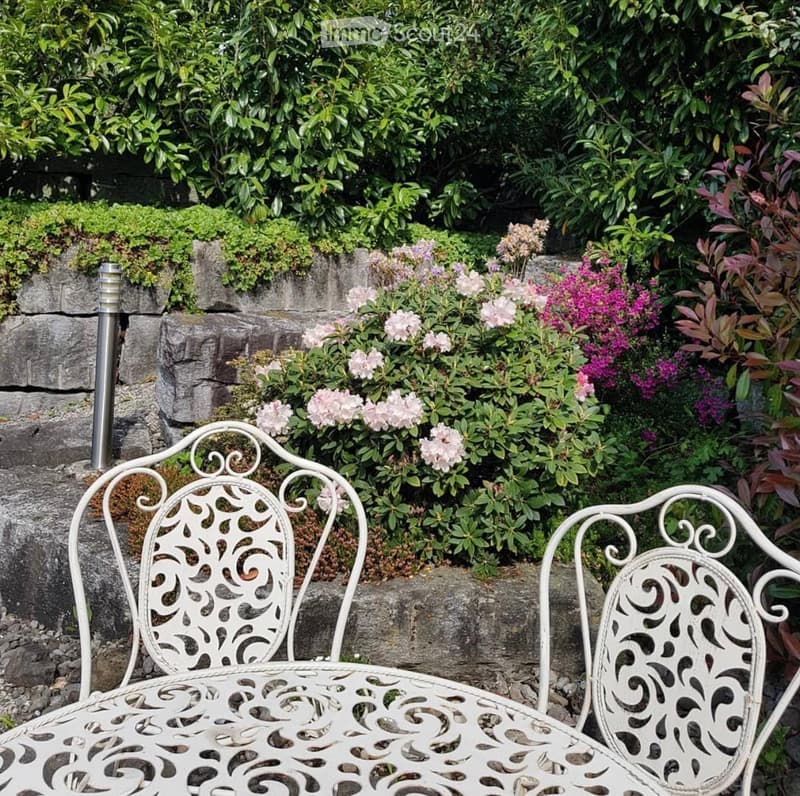 A garden area with a white metal chair and table set against a backdrop of lush greenery and flowering shrubs.