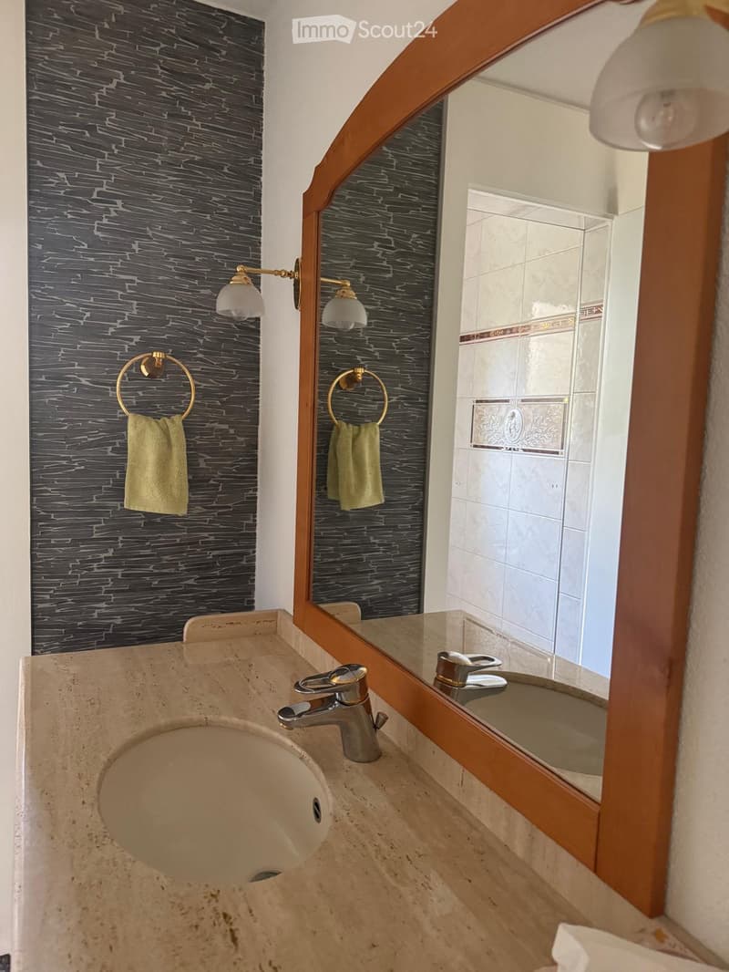 Bathroom with a sink, mirror, and towel rack. The wall has a stone-like pattern, and the mirror has a wooden frame. The sink has a faucet and a soap dish. The mirror reflects the shower area with tiled walls.