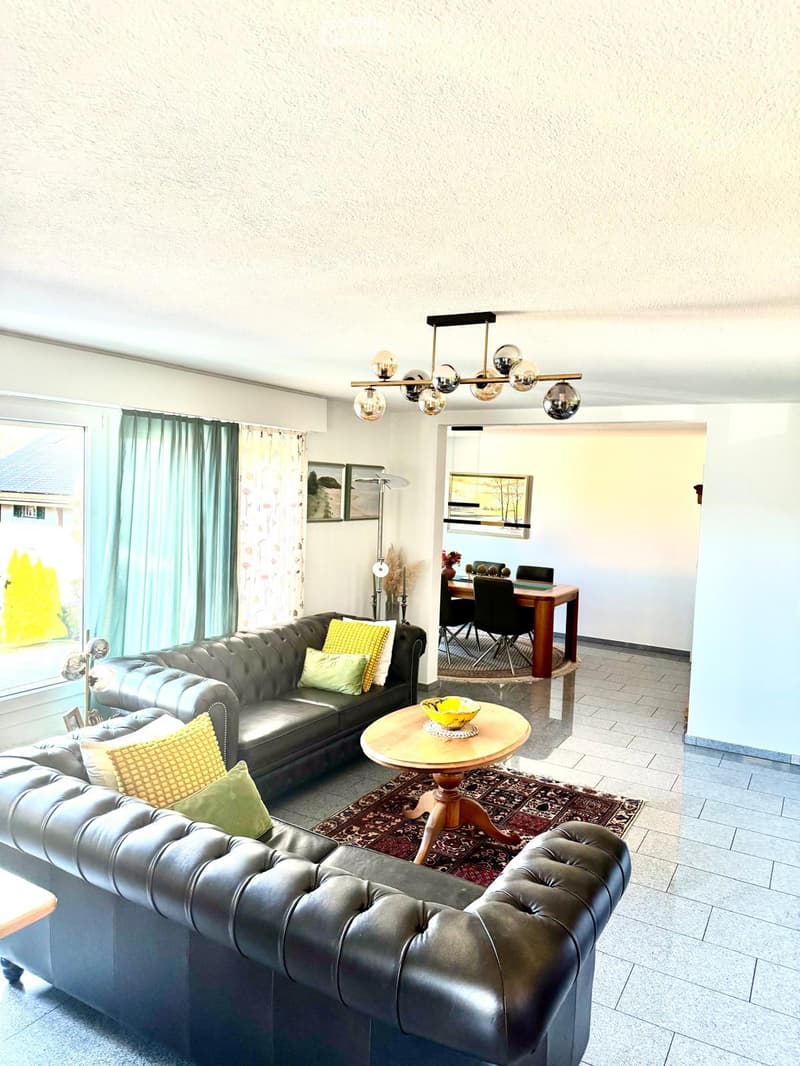 Two black couches, wooden center table, decorative items, chandelier, lamps, curtains, floor tiles, and a large window